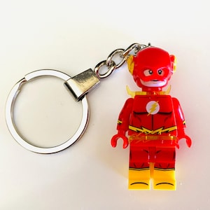 May include: A keyring featuring a red plastic minifigure character with a yellow lightning bolt emblem on the chest, yellow boots, and a red mask with yellow ear accents, attached to a circular silver metal split ring by a short chain.