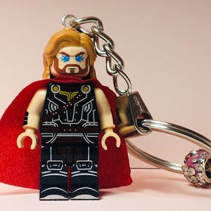 Puede incluir: Un llavero de Lego Thor con una minifigura con capa roja, pelo rubio y un disfraz detallado. El llavero incluye una cadena plateada, una anilla y un charm rosa y plateado. La figura mide aproximadamente 5 cm de alto.