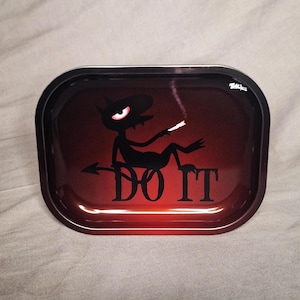 Puede incluir: Una bandeja de metal negra con un fondo degradado rojo. Un diablo de dibujos animados con un ojo rojo se representa fumando un cigarrillo y sentado en una superficie roja con el texto "DO IT" en negro.