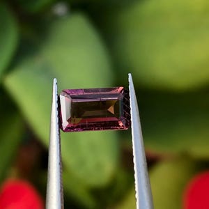 May include: A rectangular, faceted gemstone with a deep purple hue and a slight green tint. The gemstone is held in tweezers against a blurred green background.