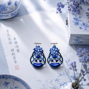 Puede incluir: Un par de pendientes colgantes con intrincados bordados de hilo azul y blanco en forma de flor, acentuados con pequeñas cuentas azules y una piedra azul lisa más grande en la base, suspendidos de ganchos de metal dorado.