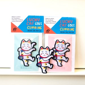 Pode incluir: Três patches de ferro com um gato da sorte de desenho animado em uma pose de escalada. Os patches estão em um fundo azul e rosa com o texto "Lucky Cat Goes Climbing Iron On Patch".