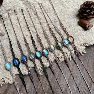 May include: A selection of macramé bracelets, each featuring an oval gemstone with iridescent qualities. The bracelets are in shades of brown and black, with gemstones displaying blue, green, and yellow tones. They are arranged on a textured, neutral-toned surface.