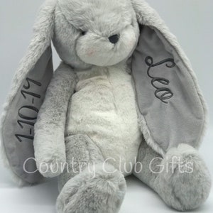 May include: A plush, light gray bunny rabbit with long, floppy ears. The bunny has a soft, fluffy texture, with a lighter gray belly and pink accents on the cheeks. The left ear has the embroidered date "1-10-19", and the right ear has the name "Leo".