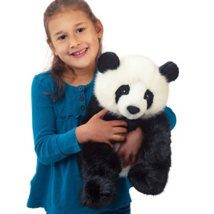 May include: A plush panda bear with black and white fur is held by a person wearing a teal long-sleeved shirt. The panda has black ears and eyes, and a white face. The stuffed animal is soft and cuddly.