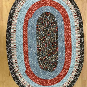 May include: Oval braided rug with a multi-colored design. The rug features concentric oval patterns in various colors, including blue, red, and brown. The outer border is dark with a paw print pattern.