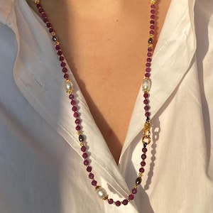 Op de afbeelding: Een lange ketting met kleine, ronde, dieprode kralen, goudkleurige accenten en grote, glanzende witte parels. De ketting wordt getoond tegen een wit overhemd, wat de kleur en het ontwerp benadrukt. Het is een sieraad.