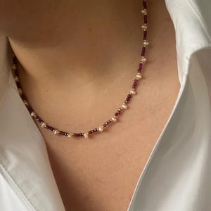 May include: A necklace with small pearls, deep red beads, and gold-coloured accents. The necklace is shown against a white collared shirt, emphasising the contrast of colours and textures. A simple and elegant piece of jewellery.