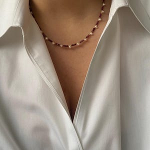 以下が含まれることがあります： 小さな丸い赤いビーズと白い真珠が交互に並んだ繊細なネックレス。白い襟付きシャツの上に着用されています。
