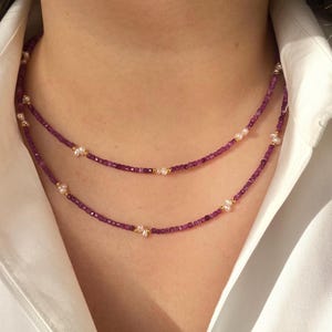 Op de afbeelding: Twee lagen ketting met kleine, dieppaarse kralen, witte parels en goudkleurige accenten. De ketting wordt getoond tegen een wit overhemd.