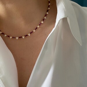 以下が含まれることがあります： 小さな丸い真珠と深紅の宝石が特徴の繊細なネックレス。金色のビーズが散りばめられています。白い襟付きシャツの上から着用されています。