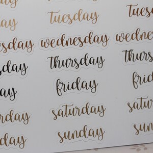 May include: A sheet of stickers with the days of the week written in a cursive font. The stickers are white with gold and black lettering.