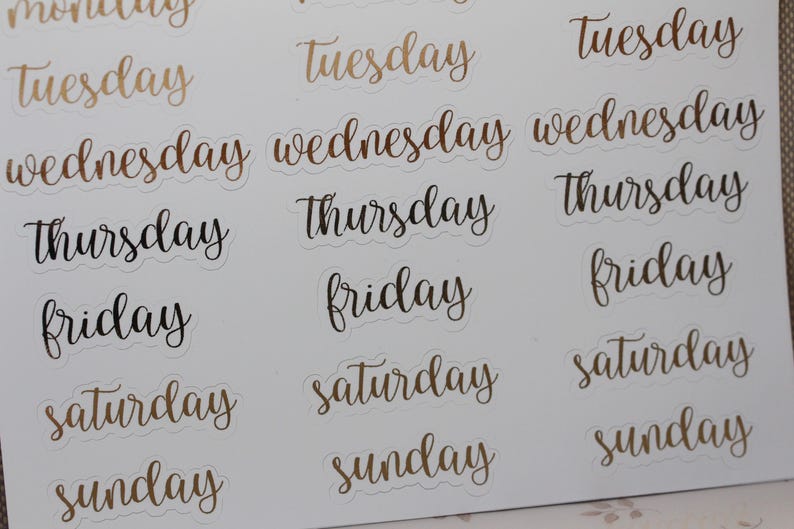 May include: A sheet of stickers with the days of the week written in a cursive font. The stickers are white with gold and black lettering.
