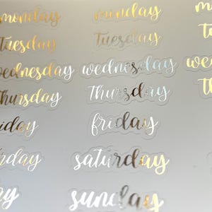 May include: A sheet of clear stickers with gold and silver foil lettering. The stickers feature the days of the week: Monday, Tuesday, Wednesday, Thursday, Friday, Saturday, and Sunday.