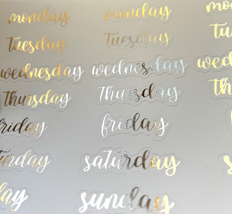 May include: A sheet of clear stickers with gold and silver foil lettering. The stickers feature the days of the week: Monday, Tuesday, Wednesday, Thursday, Friday, Saturday, and Sunday.