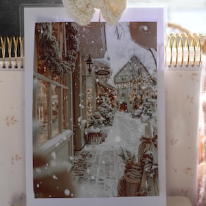 May include: A winter scene print featuring a snow-covered street with shops and festive decorations. The image is held by a gold spiral clip and a cream-colored bow. The print is displayed in a clear holder with a floral design.