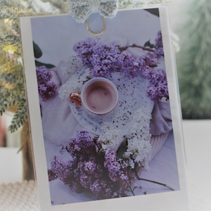 Può includere: Primo piano di una tazza da tè in porcellana bianca con un motivo floreale rosa, riempita di una bevanda rosa chiaro. La tazza da tè è circondata da un pizzo bianco e fiori di lillà viola.