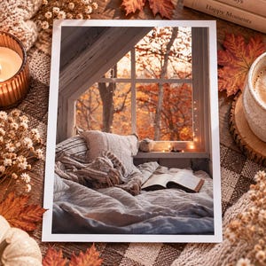 May include: A rectangular art print featuring a cozy window nook scene with a soft bed, a textured throw blanket, and an open book. The window overlooks a vibrant autumn forest with orange leaves. The print is framed by a white border on a rustic surface.