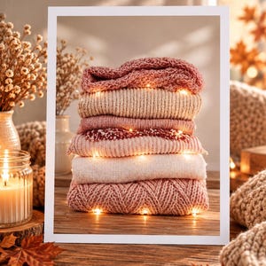 Könnte beinhalten: Ein Stapel aus fünf gefalteten Strickpullovern in Rosa-, Creme- und Beigetönen, beleuchtet durch kleine warmweiße Lichterketten, die zwischen den Schichten liegen. Die Pullover weisen verschiedene Texturen wie Ripp-, Zopf- und Grobstrickmuster auf.