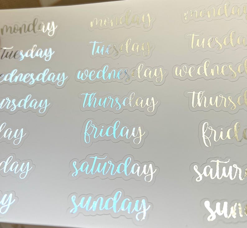May include: A sheet of clear stickers with the days of the week written in a cursive font. The stickers are iridescent and have a holographic finish. The days of the week are: Monday, Tuesday, Wednesday, Thursday, Friday, Saturday, and Sunday.