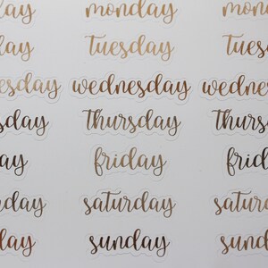 May include: A sheet of gold foil stickers with the days of the week written in a cursive font. The stickers are on a white background.