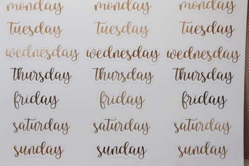 May include: A sheet of gold foil stickers with the days of the week written in a cursive font. The stickers are on a white background.