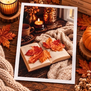 Peut inclure: Une impression rectangulaire montrant un livre ouvert avec deux feuilles d'érable orange et rouge sur les pages, entouré d'une couverture en tricot beige, d'une bougie allumée, d'une tasse brune et d'une petite citrouille sur une surface en bois.