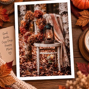 May include: A white wooden chair sits in a forest setting with a wreath of dried orange flowers, a lit metal lantern, and a draped beige throw blanket. A card nearby reads: Good Books, Cozy Days, Happy Plans, with a heart symbol drawn below.