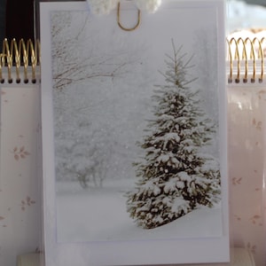 May include: A winter-themed desk calendar featuring a snowy landscape with a snow-covered evergreen tree. The calendar has a gold spiral binding and a decorative clip. The background includes a woven basket and a pink knitted item.