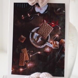 以下が含まれることがあります： ホットチョコレートとマシュマロで満たされたマグカップのクローズアップ。グラハムクラッカーとストリングライトに囲まれています。シーンは、素朴な雰囲気のある暗い木製の表面に設定されています。