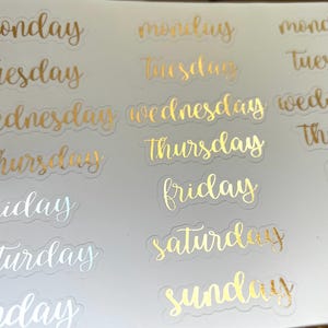 May include: Gold foil stickers with the days of the week written in a cursive font. The stickers are on a white background.
