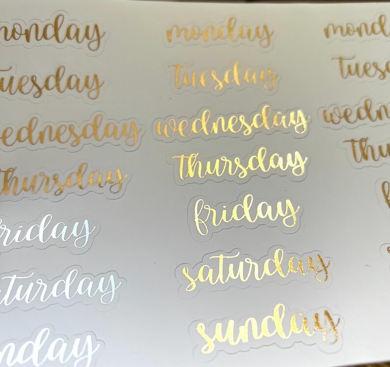 May include: Gold foil stickers with the days of the week written in a cursive font. The stickers are on a white background.