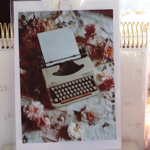 Könnte beinhalten: Eine cremefarbene Vintage-Schreibmaschine mit schwarzen Tasten liegt auf einer floral gemusterten Oberfläche. Das Bild wird von einem transparenten Acrylhalter mit goldenen Akzenten umrahmt. Weiche rosa und weiße Blumen sind verstreut.