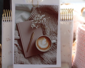 Pode incluir: Imagem estética com um caderno rosa, caneta dourada e uma xícara de arte latte em um cobertor aconchegante. Flores brancas delicadas e um caderno espiral dourado contribuem para a cena serena e convidativa. O tema geral é relaxamento e conforto.