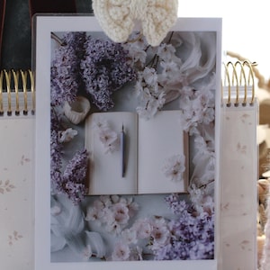 May include: A photograph of an open book with a pen, surrounded by lilac and white flowers. The image is displayed in a clear frame, with a cream-colored spiral notebook on either side. A crocheted bow is at the top.