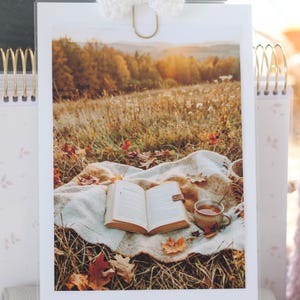 Könnte beinhalten: Ein vertikaler rechteckiger Druck mit einem aufgeschlagenen Buch und einer Keramiktasse auf einer cremefarbenen Decke in einem herbstlichen Grasfeld. Die Szene zeigt gefallene orangefarbene und braune Blätter bei goldenem Sonnenuntergang.