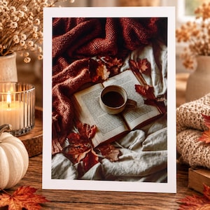 Peut inclure: Une impression rectangulaire montrant un livre ouvert posé sur un tissu blanc, surmonté d'une petite tasse en céramique remplie d'un liquide sombre. Une couverture en tricot rouille texturée et des feuilles d'automne séchées entourent le livre.