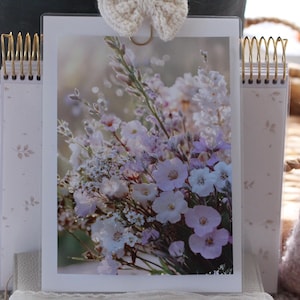 Könnte beinhalten: Gerahmtes Bild mit Blumendruck von weißen und lila Blumen. Der Rahmen hat eine goldene Spiralbindung. Eine cremefarbene gehäkelte Schleife ist oben am Rahmen befestigt. Ein rosa Strickartikel befindet sich im Hintergrund. Das Bild ist in einem transparenten Rahmen.