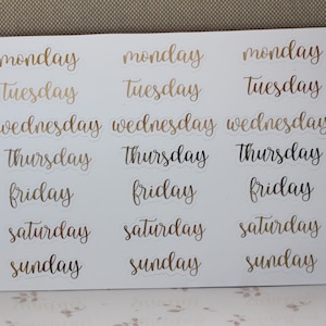 May include: A sheet of gold foil stickers with the days of the week written in cursive script. The stickers are on a white background.