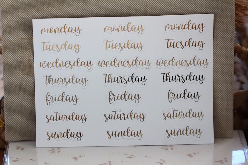 May include: A sheet of gold foil stickers with the days of the week written in cursive script. The stickers are on a white background.