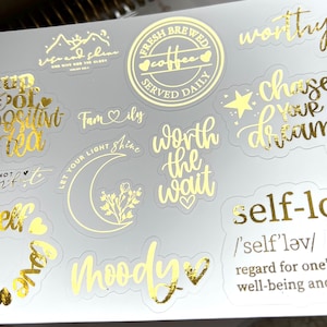 May include: A sheet of gold foil stickers with various inspirational quotes and phrases, including "cup of positive tea", "worth the wait", "self-love", "moody", "chase your dreams", and "let your light shine".