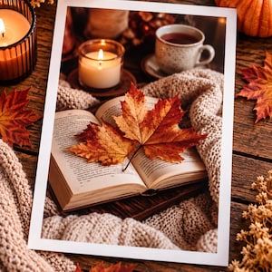 May include: A rectangular photograph print featuring an open book with two orange and brown maple leaves resting on the pages. The scene includes a lit candle in a glass holder, a ceramic mug, and a beige knit blanket on a dark wooden surface.