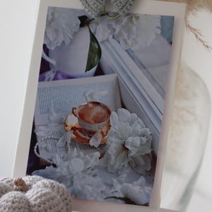 Könnte beinhalten: Eine weiße Porzellan-Teetasse mit Untertasse und einer braunen Flüssigkeit darin, umgeben von weißen Blumen und einem Buch mit einer weißen Spitzenüberlage. Das Bild ist mit einem weißen Rahmen versehen.