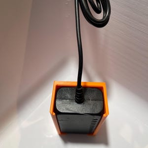 May include: A black power adapter with a black cord, mounted in an orange plastic wall mount.