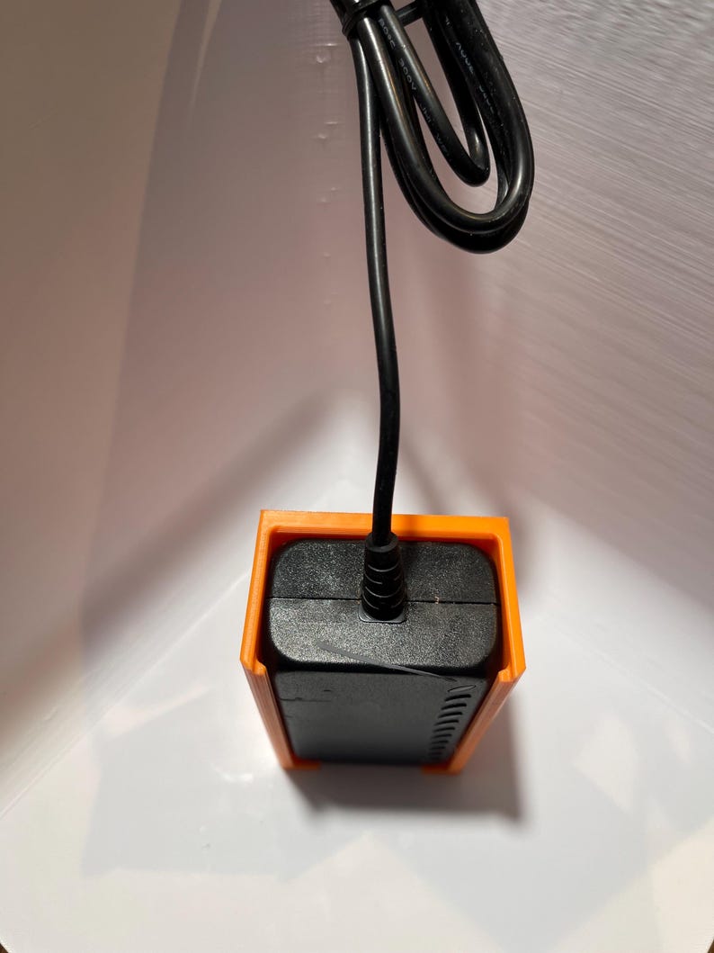 May include: A black power adapter with a black cord, mounted in an orange plastic wall mount.