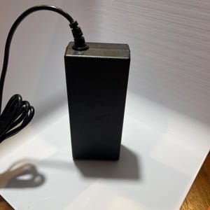 May include: A black power adapter with a black power cord. The adapter has a rectangular shape and is plugged into an electrical outlet.