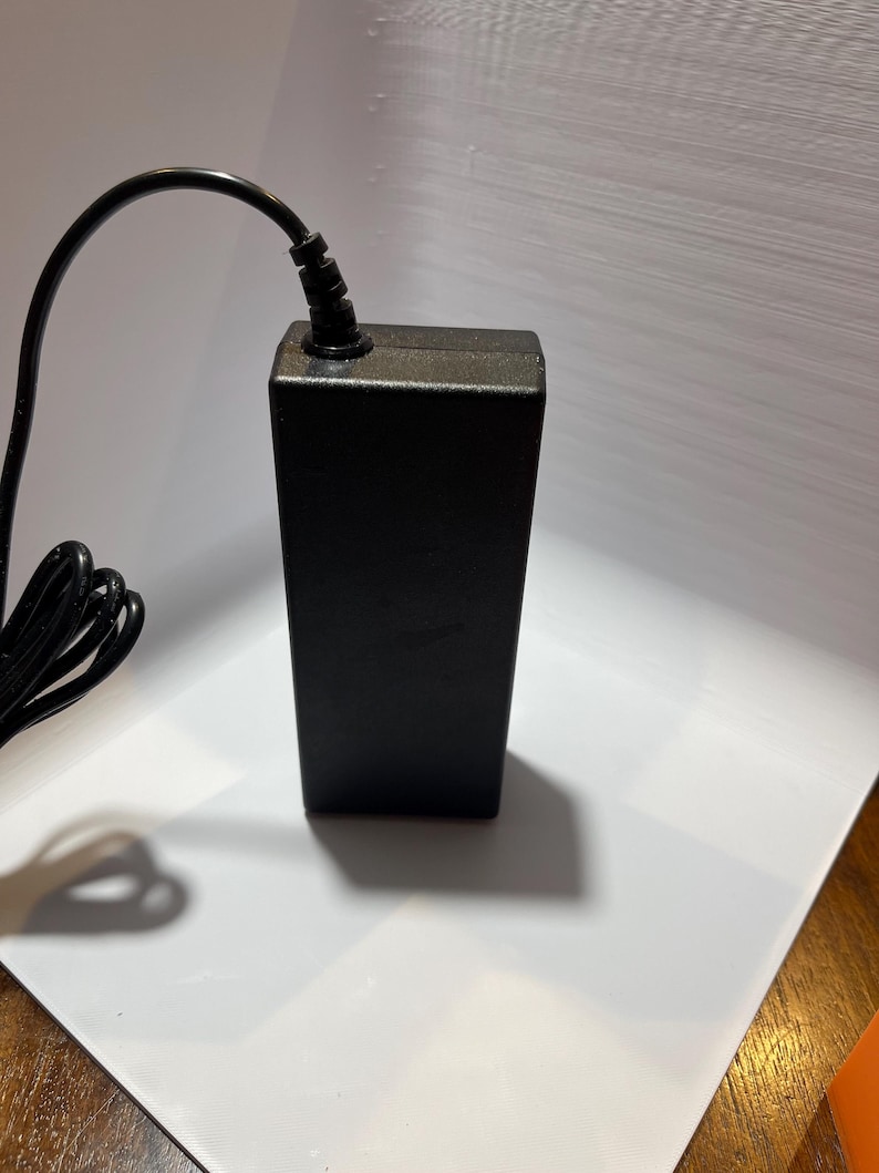 May include: A black power adapter with a black power cord. The adapter has a rectangular shape and is plugged into an electrical outlet.
