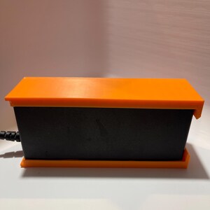 May include: A black rectangular power adapter with an orange top and bottom. The adapter has a power cord attached to it.
