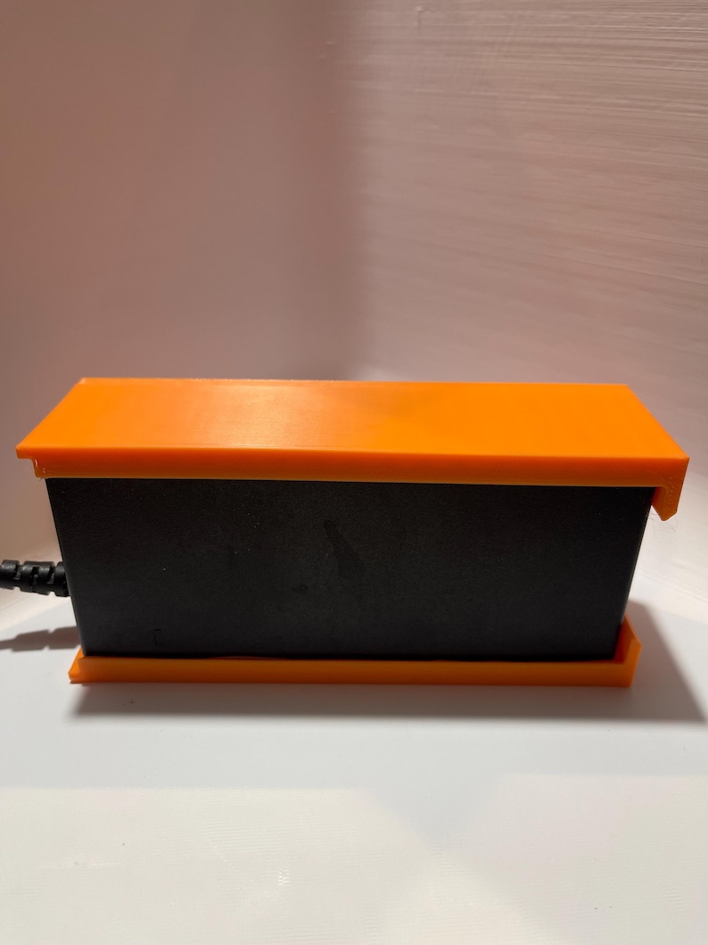 May include: A black rectangular power adapter with an orange top and bottom. The adapter has a power cord attached to it.