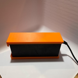 May include: A black rectangular box with an orange lid and a black cord attached to the side.